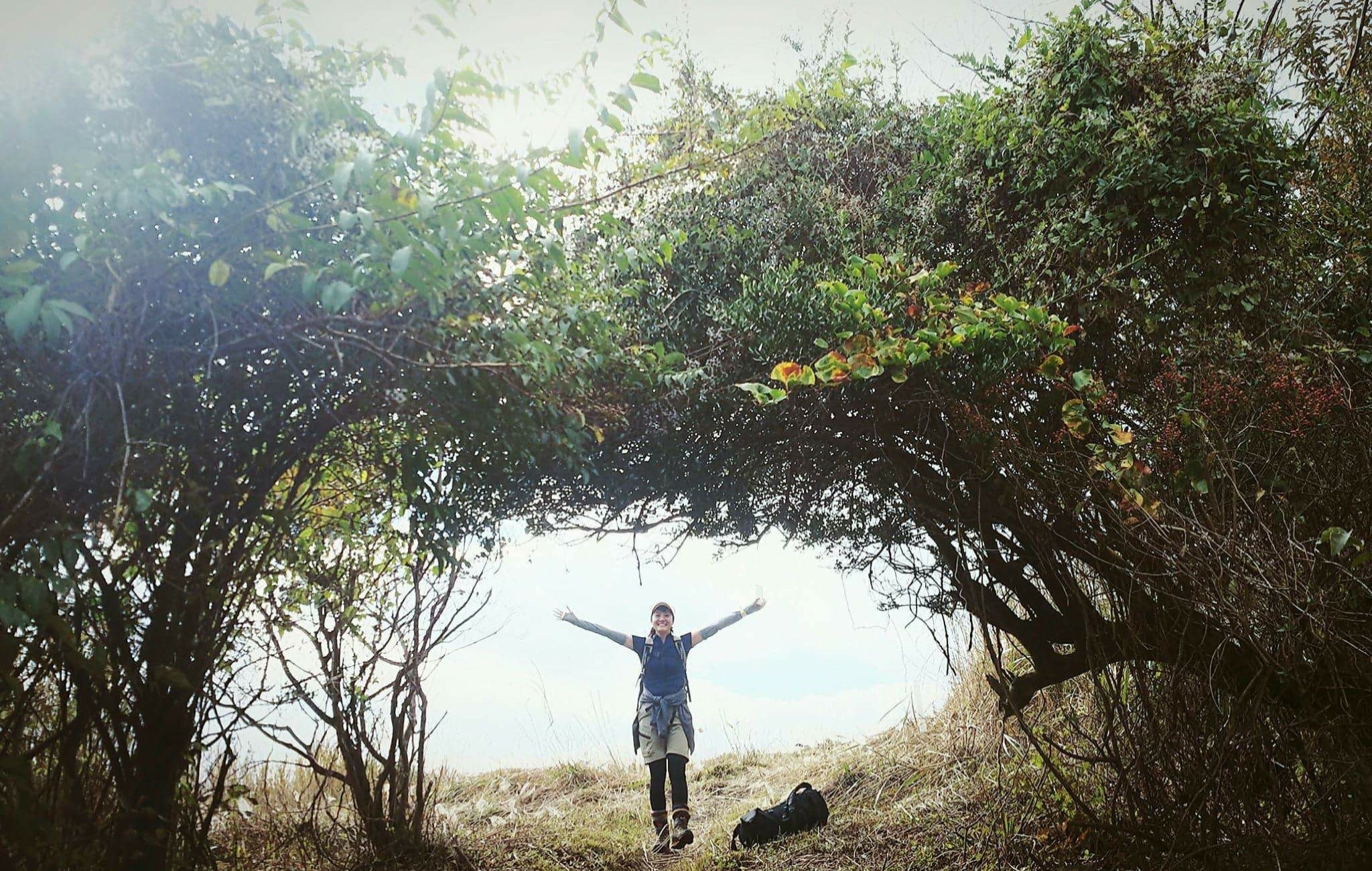 Nozomi standing with arms open under an arch of coastal trees on a bright hillside path