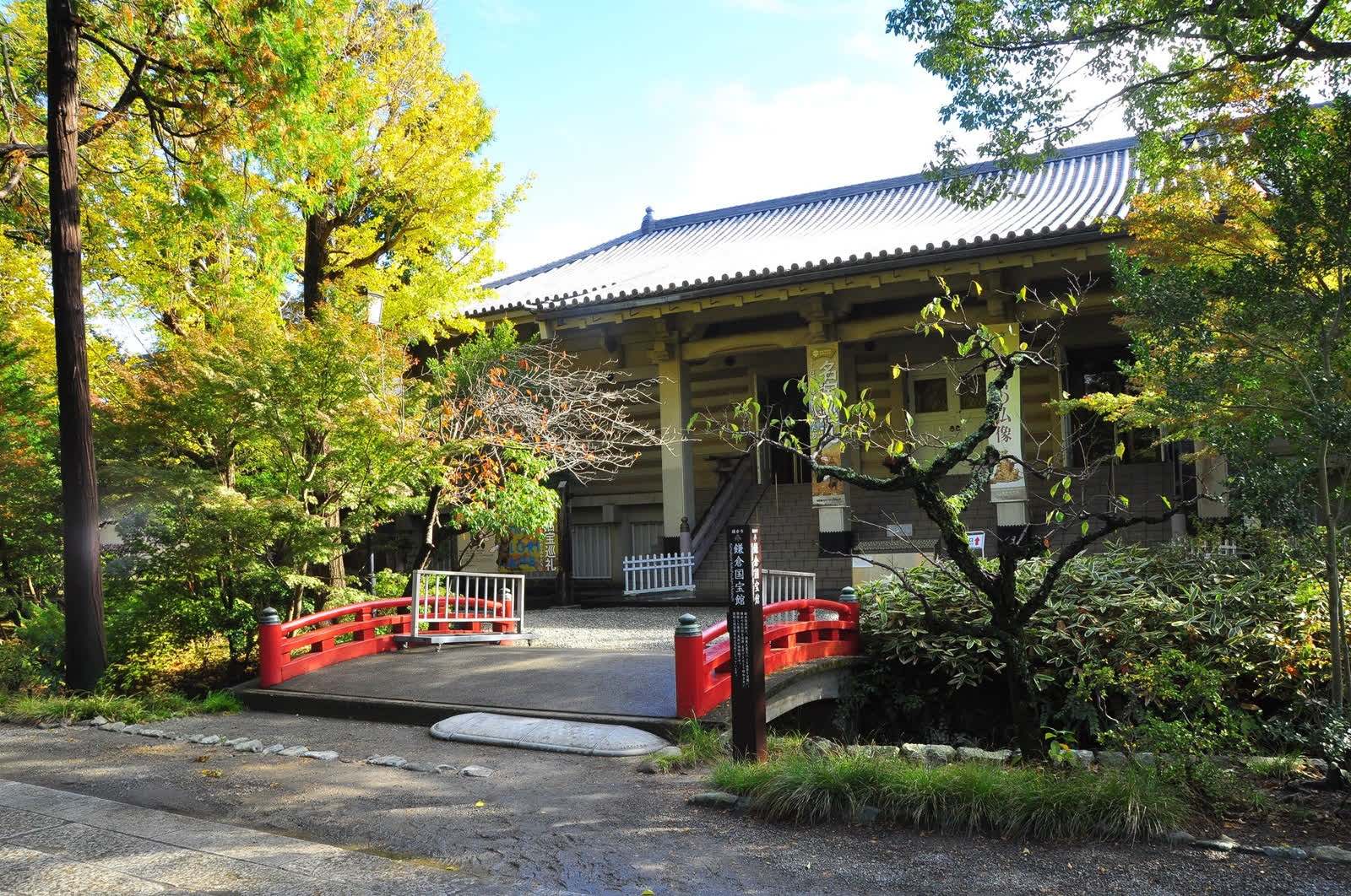 A vermilion arched bridge and the stone marker of the Kamakura Museum of National Treasures, beneath yellowing ginkgo